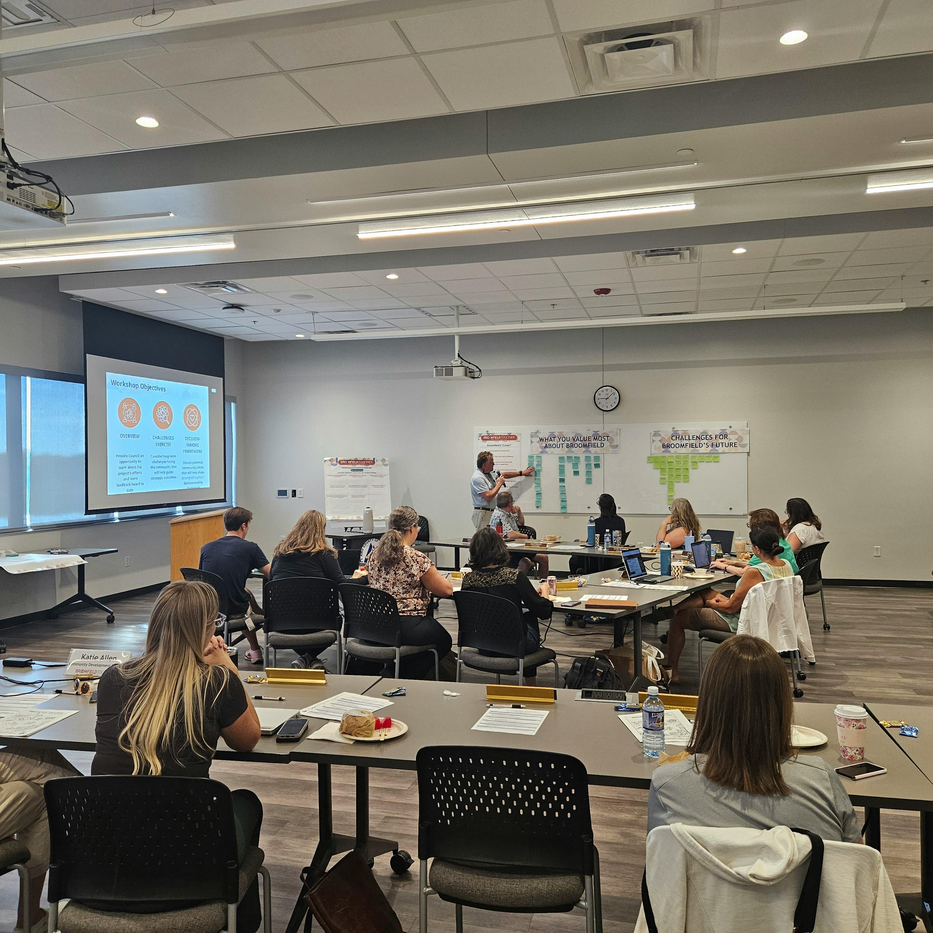 Photograph of a community workshop in a conference room. A facilitator stands at the front, pointing to a whiteboard titled "What You Value Most About Broomfield" which is covered in blue sticky notes. Participants sit at tables with laptops.