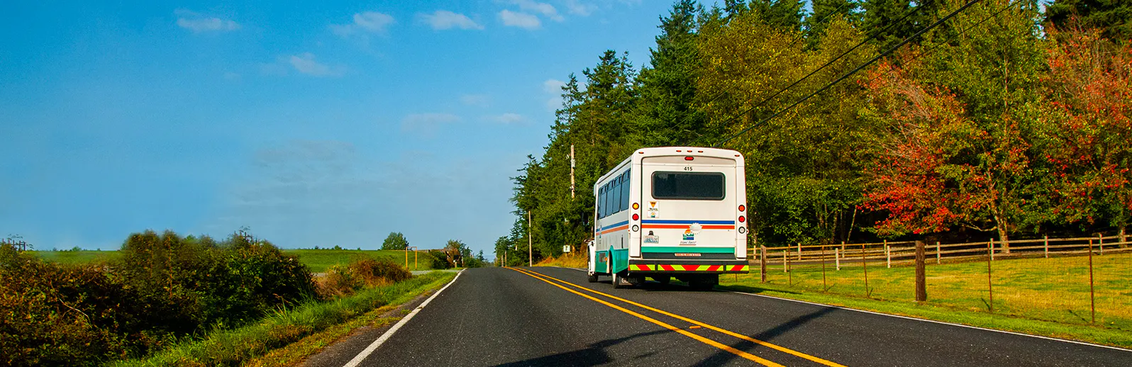 Image of bus driving down rural road with blue sky, trees and a green field 
