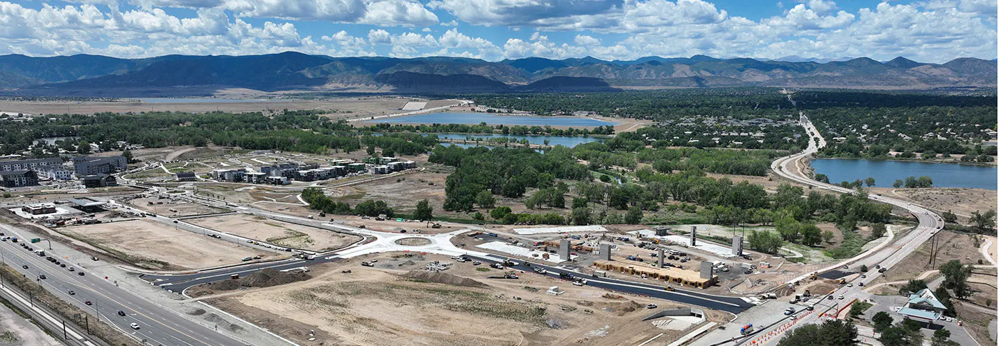 Aerial view of roundabout under construction surrounded by unfinished roads, work vehicles, buildings, mountains in distance.