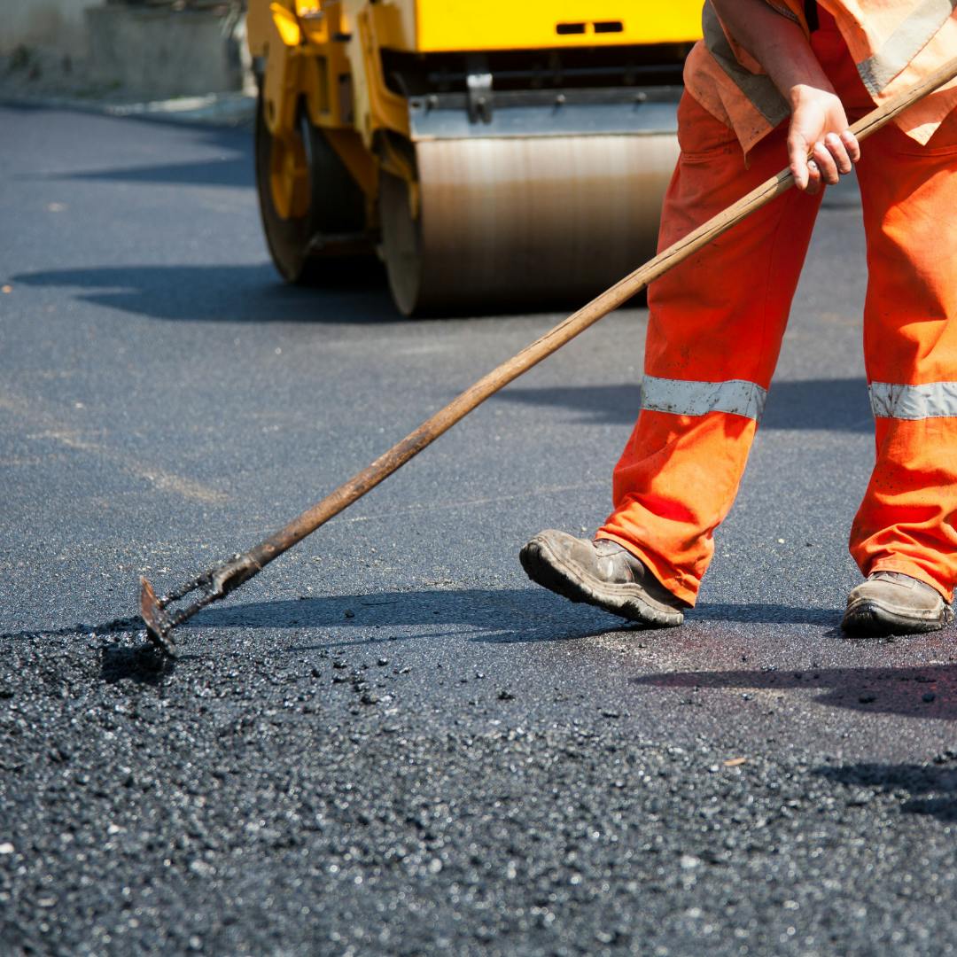 A worker in orange safety gear spreading asphalt to repair pavement.