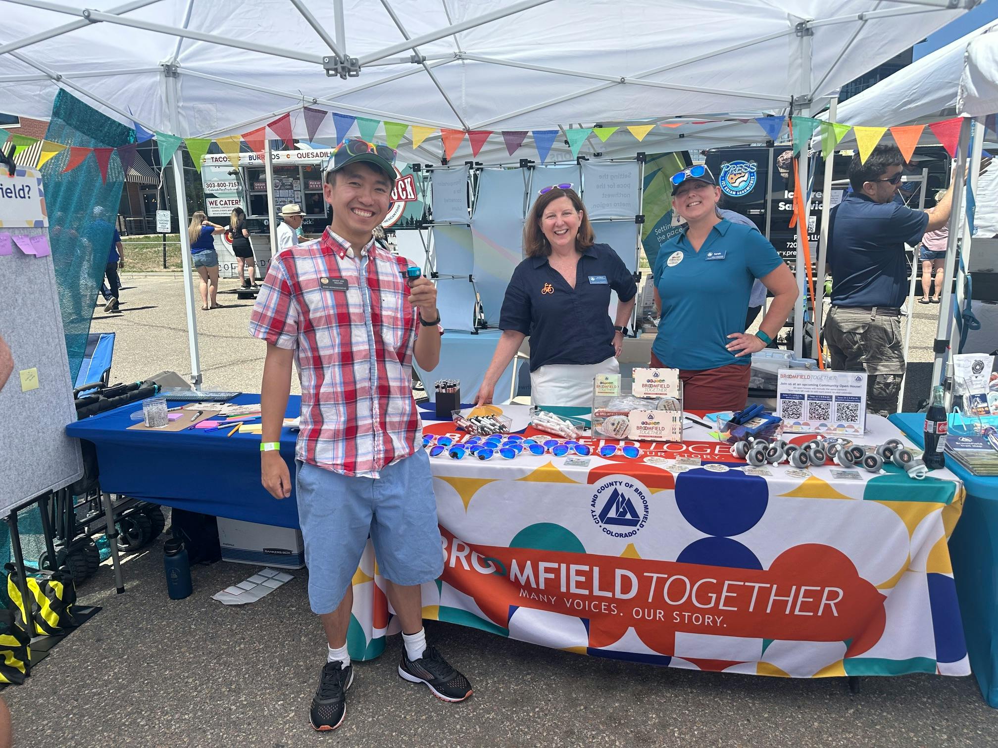 Representative Kenny Nguyen stands in a red plaid shirt and blue shorts smiling for the camera. He's standing in front of a table with a colorful Broomfield Together tablecloth. Two women staffing the booth stand behind the table smiling for the camera.