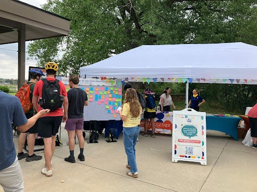 Photograph of a community engagement event outdoors under a white tent. People are gathered around a large board covered in colorful sticky notes. A white A-frame sign in the foreground reads, "What do you love about Broomfield?”