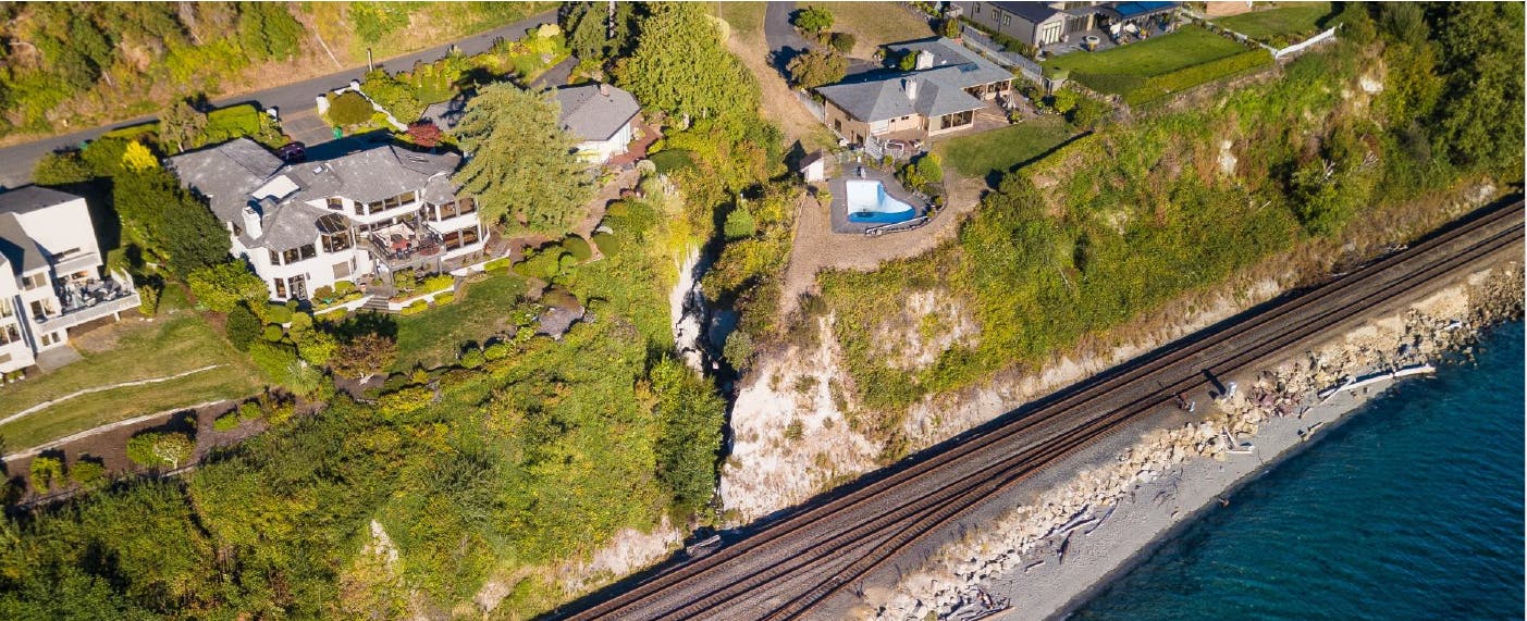 Aerial image of the area showing a large eroded area near the train tracks and extending upwards between two homes