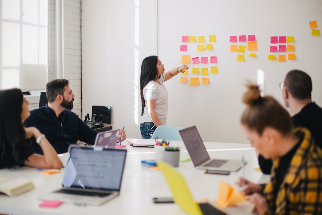 people sitting at table with laptops for a brainstorming meeting  with one person standing with sticky notes