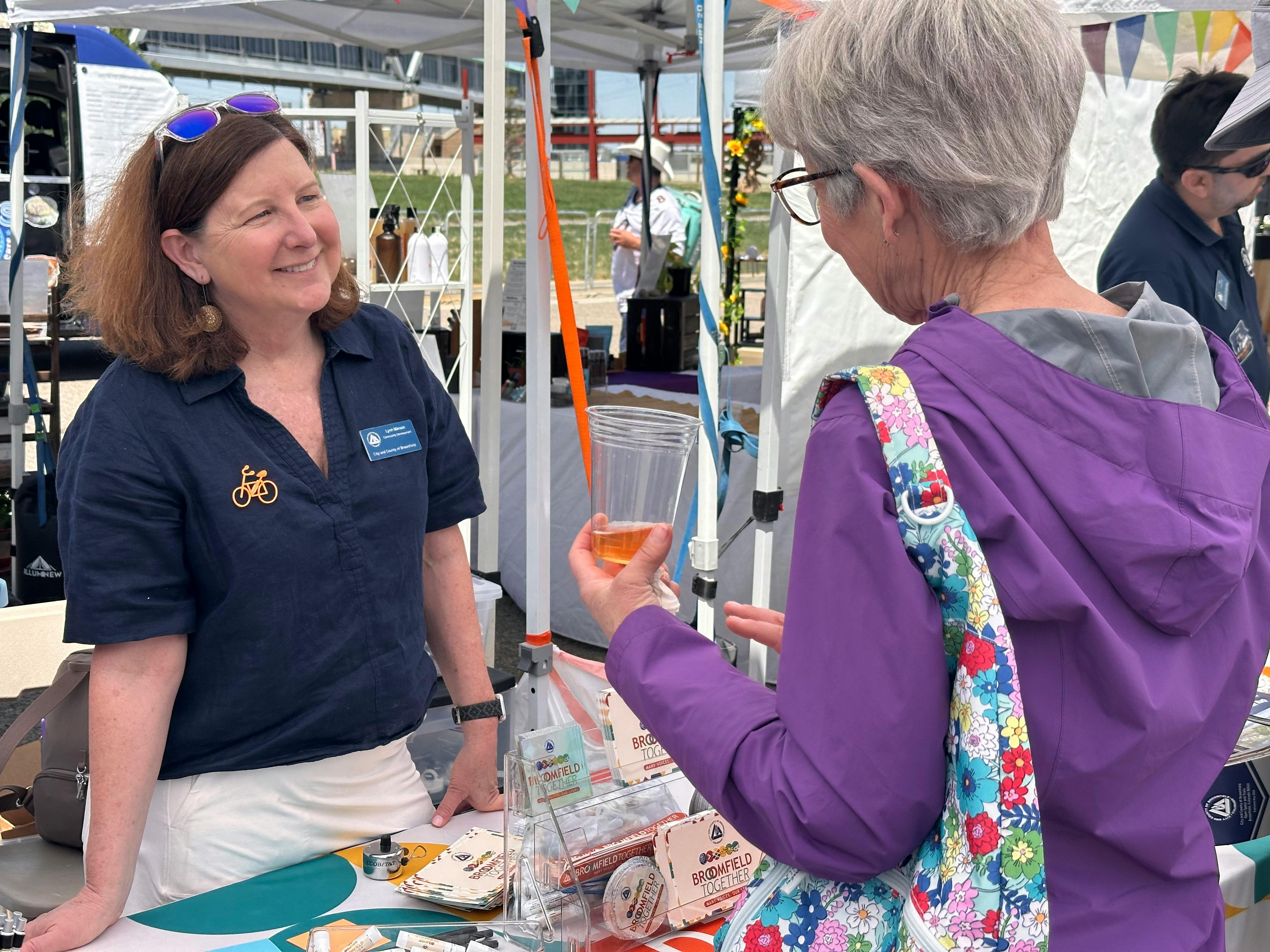 A woman wearing a City and County of Broomfield name tag and a bicycle pin smiles while interacting with a visitor at an outdoor event booth. The table in front of her displays "Broomfield Together" brochures and promotional materials.