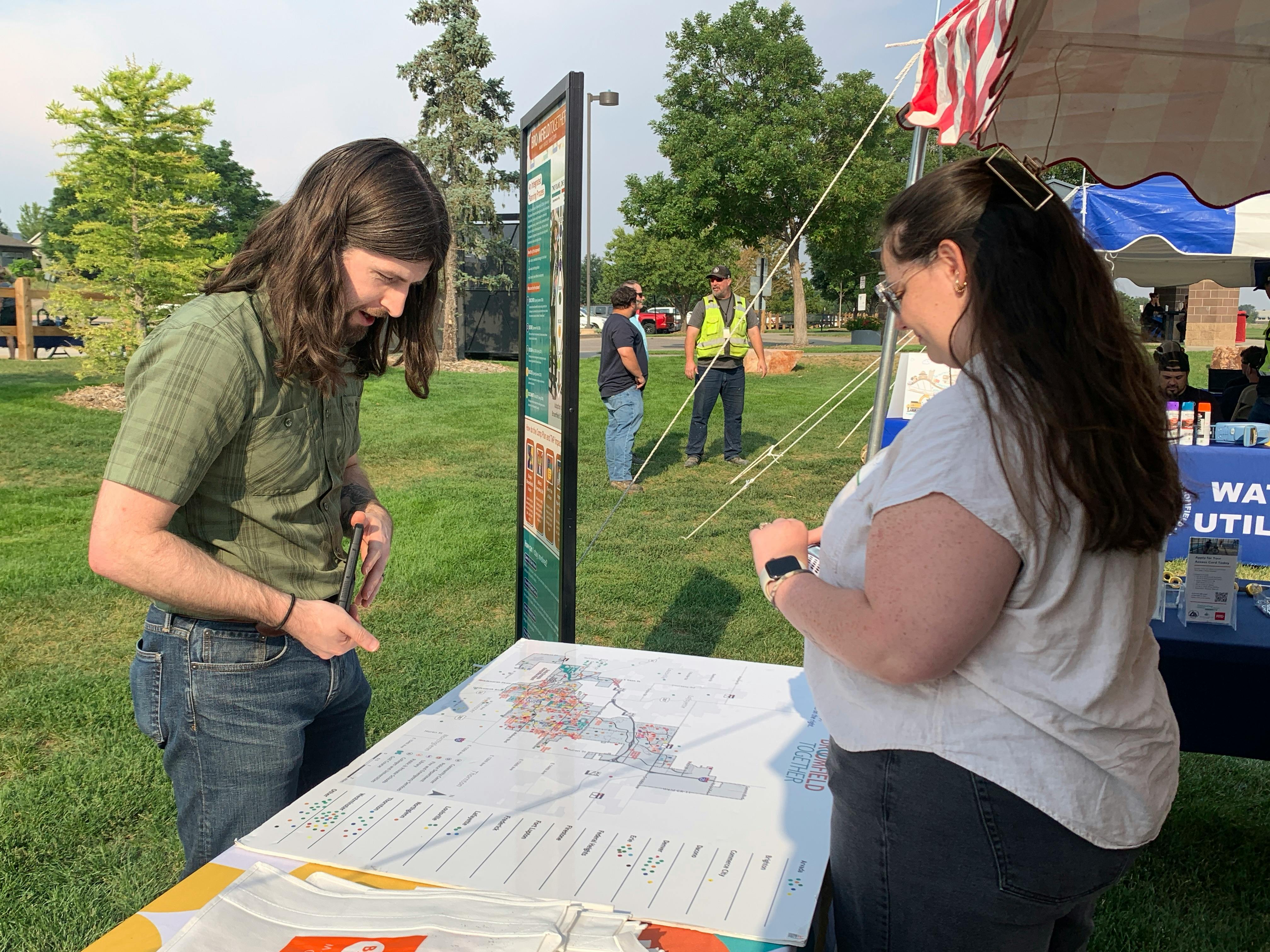 A man and a woman look at a large map titled "Broomfield Together" on a table at an outdoor community event. The man leans over the map while the woman stands opposite him. In the background, there are other people on a grassy field.