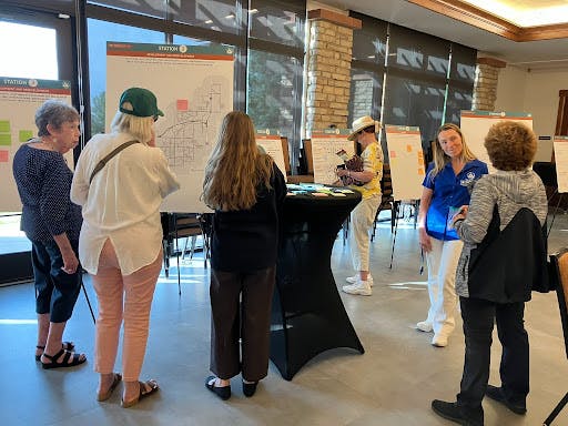 A community engagement workshop where participants are interacting with informational posters on easels. People are standing around the posters and a central table with sticky notes, discussing and providing feedback on local planning initiatives.