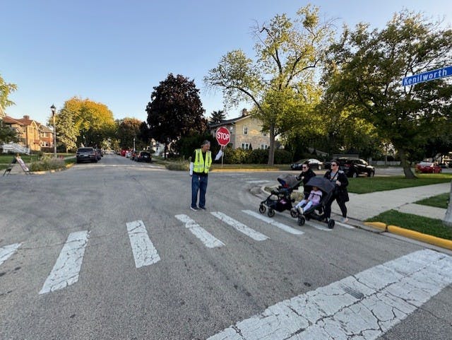 Two people walking with strollers as a crossing guard watches on.