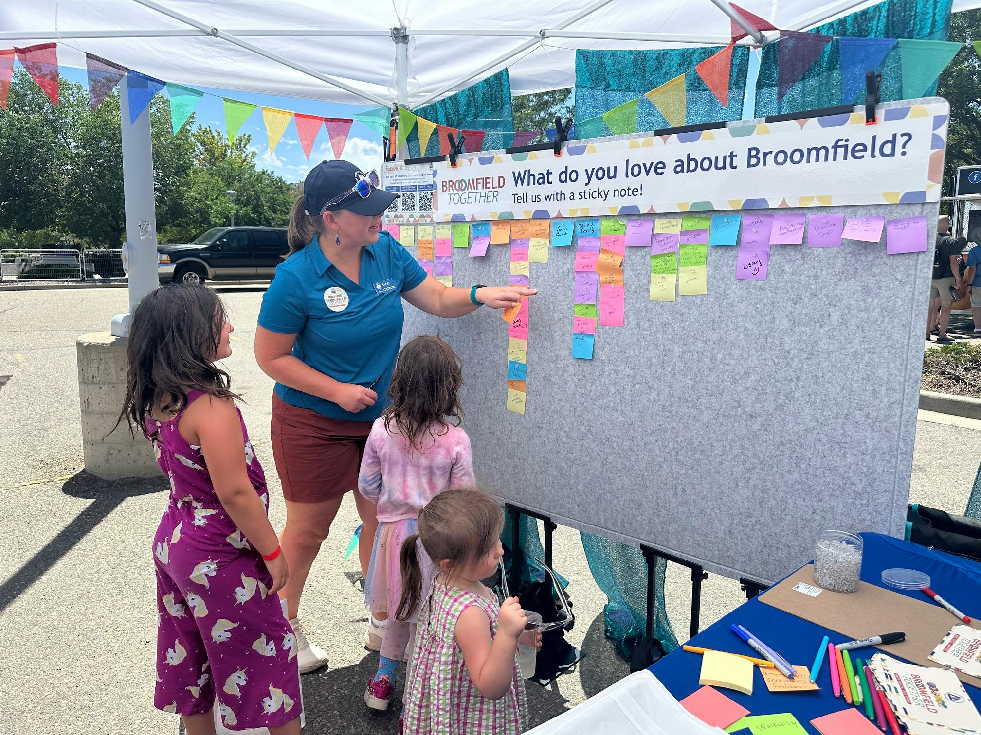 A woman in a blue polo shirt and baseball cap points to a sticky note on a large gray display board while three young girls look on. The board is covered with many colorful sticky notes containing handwritten feedback. 