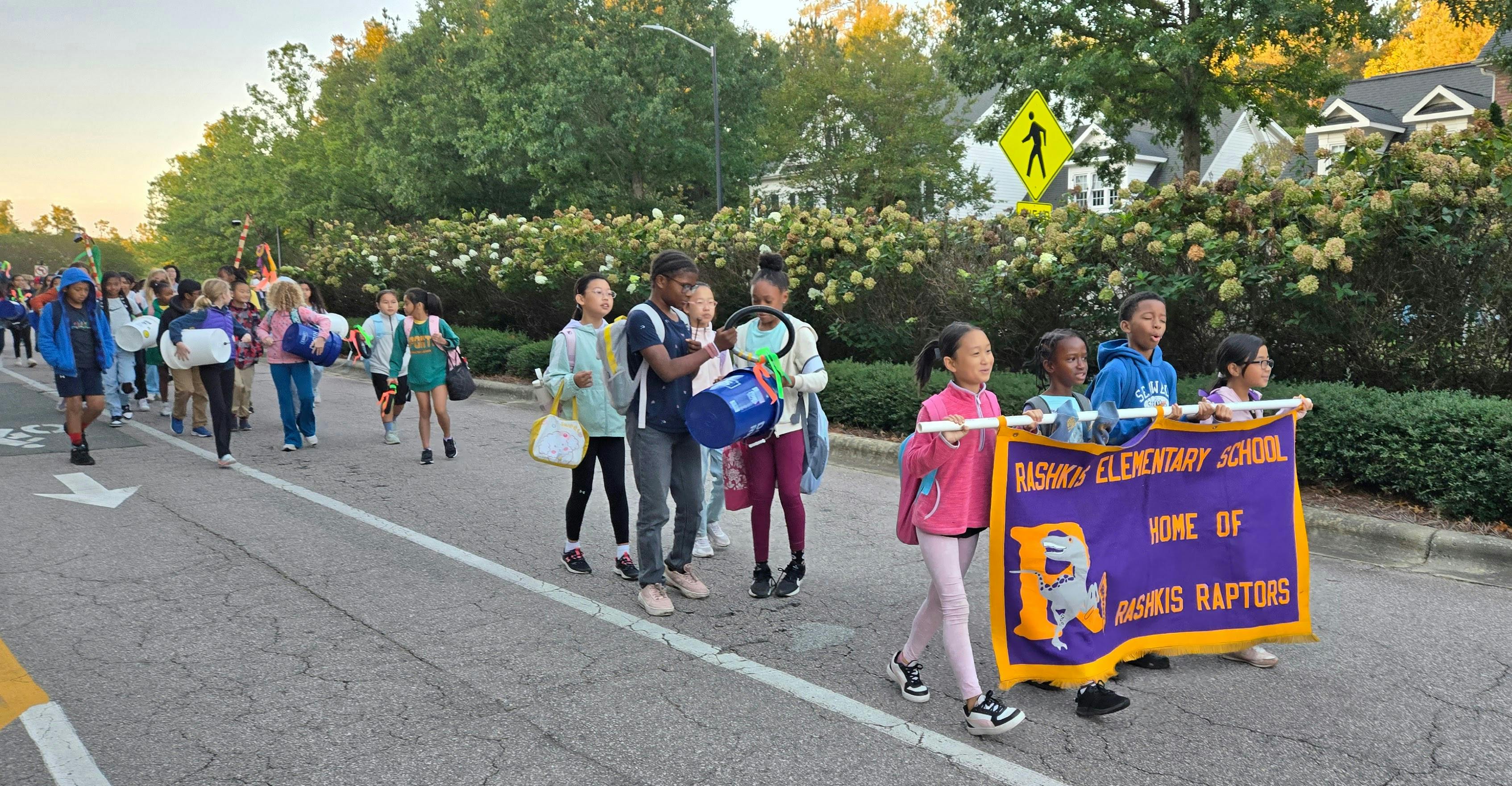 Kids and adults walk to school with a banner that says "Rashkis Elementary School Home of the Rashkis Raptors"