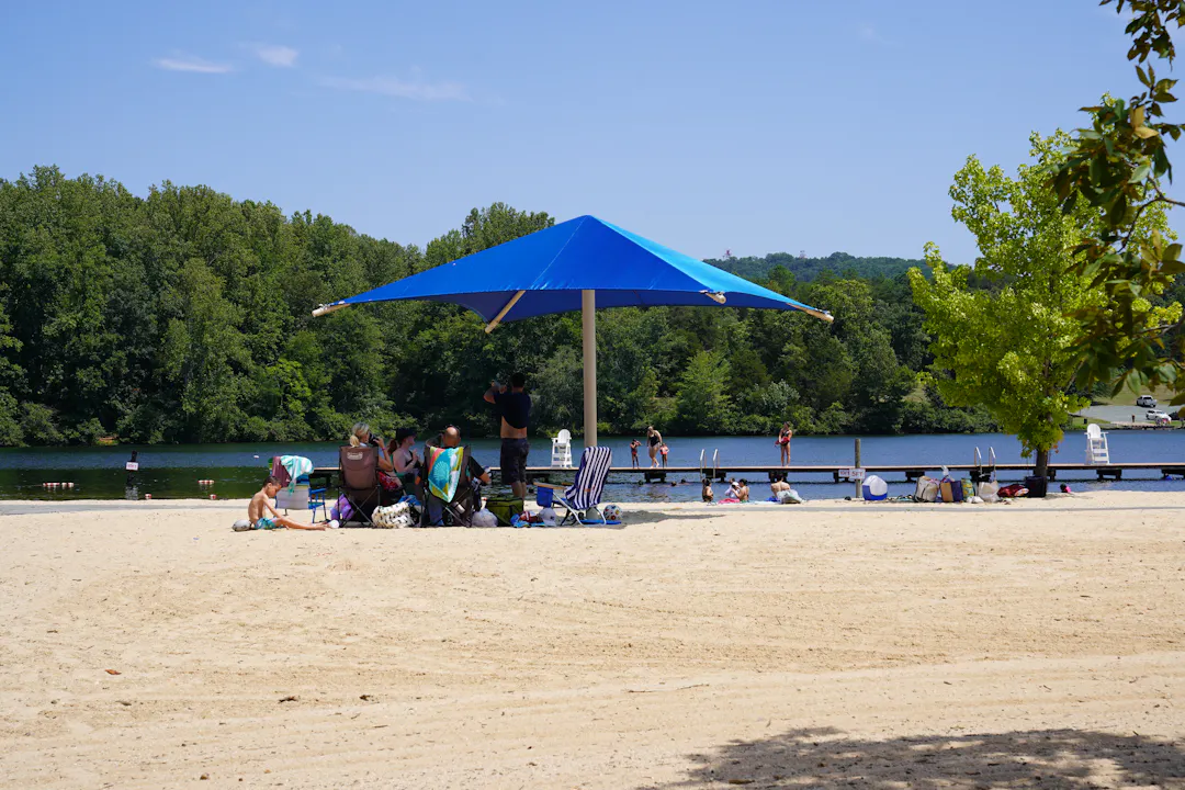 A sunny day at Chris Greene Lake with people relaxing under blue sunshade in beach chairs in sand, next to swimming shoreline