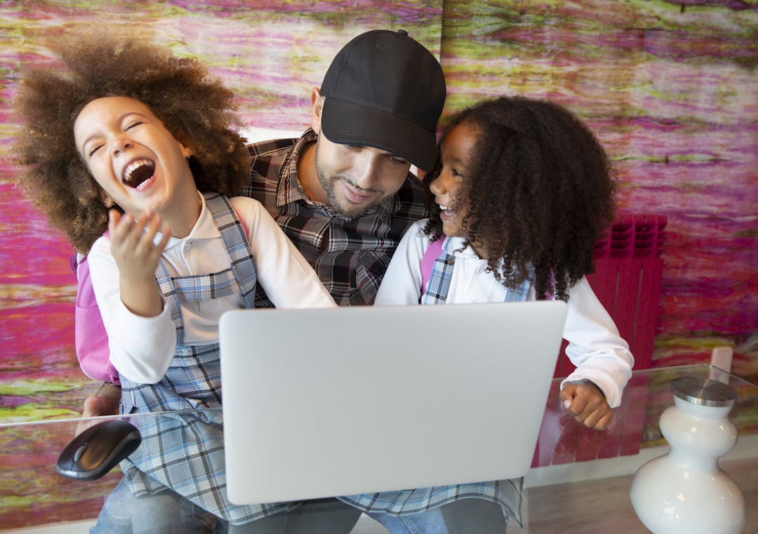 Dad looking at the internet on a computer while his two daughters laugh. 