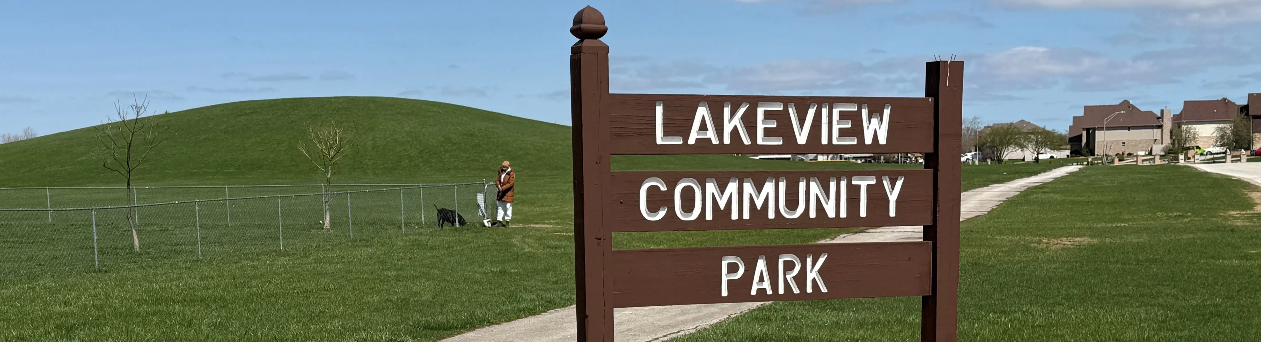 Sign for Lakeview Community Park, with a person and a dog near the fenced in dog park in the background. 
