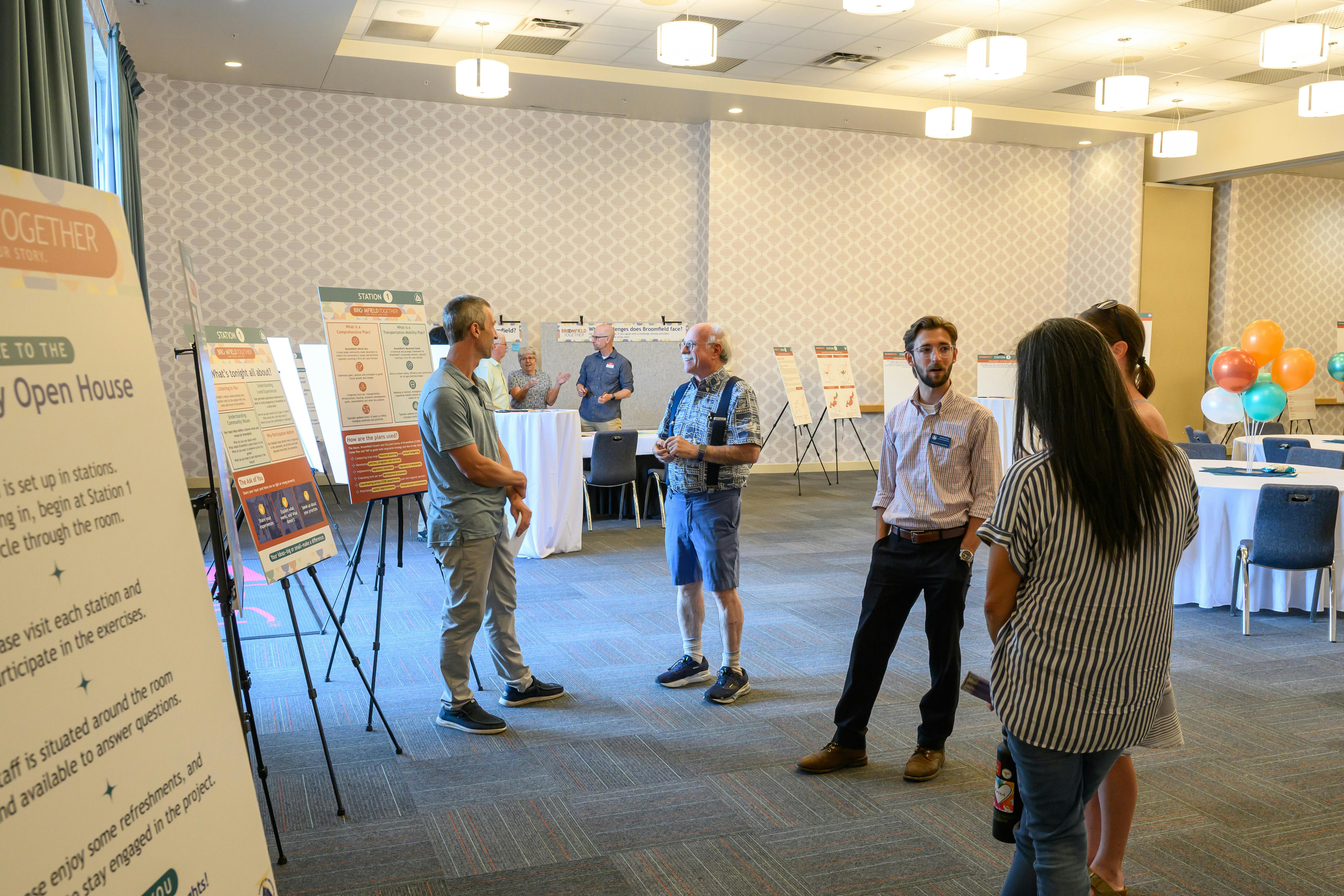 A photograph of a community open house event in a large, well-lit room. Several people are engaged in conversation near informational posters mounted on easels.