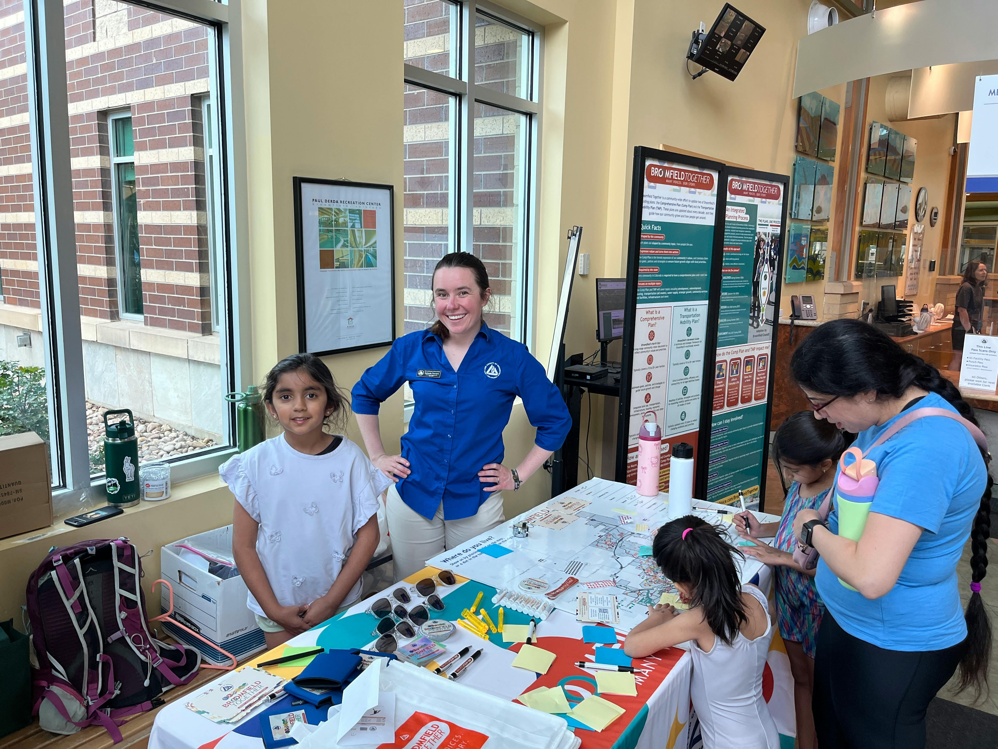 A woman in a blue shirt and a young girl stand behind a community engagement table at an indoor event. Other children and an adult are interacting with a map and sticky notes on the table.