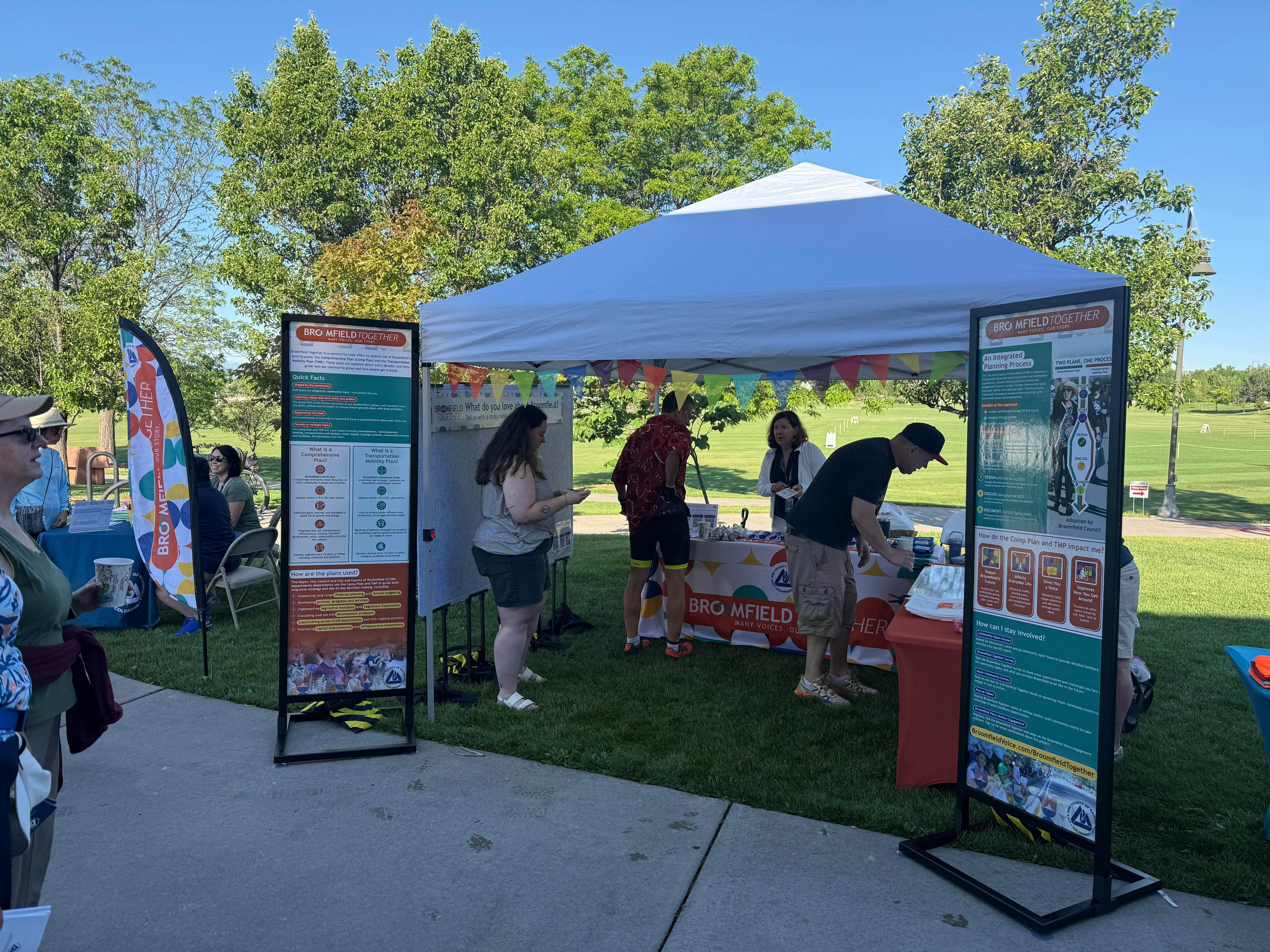 Four people stand under a white canopy tent looking at images on tables. Colorful flags hang from the tent canopy and two large colorful signs frame either side of the tent entrance.