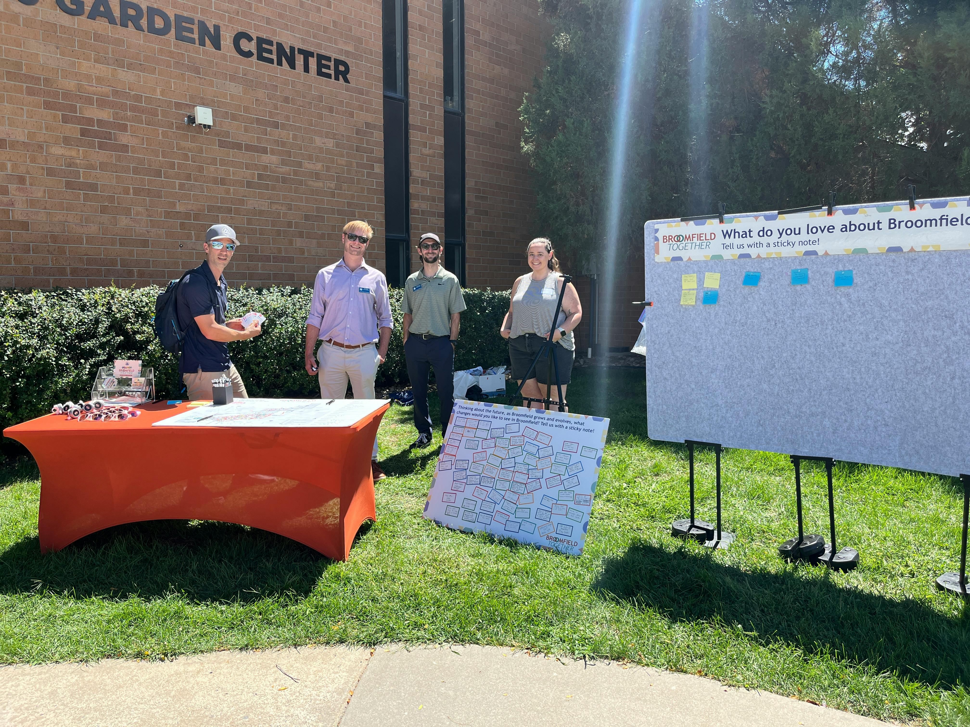 Photograph of four people at an outdoor community engagement booth for "Broomfield Together" in front of a brick building. A man stands behind an orange table with materials, while three others stand nearby on the grass. 