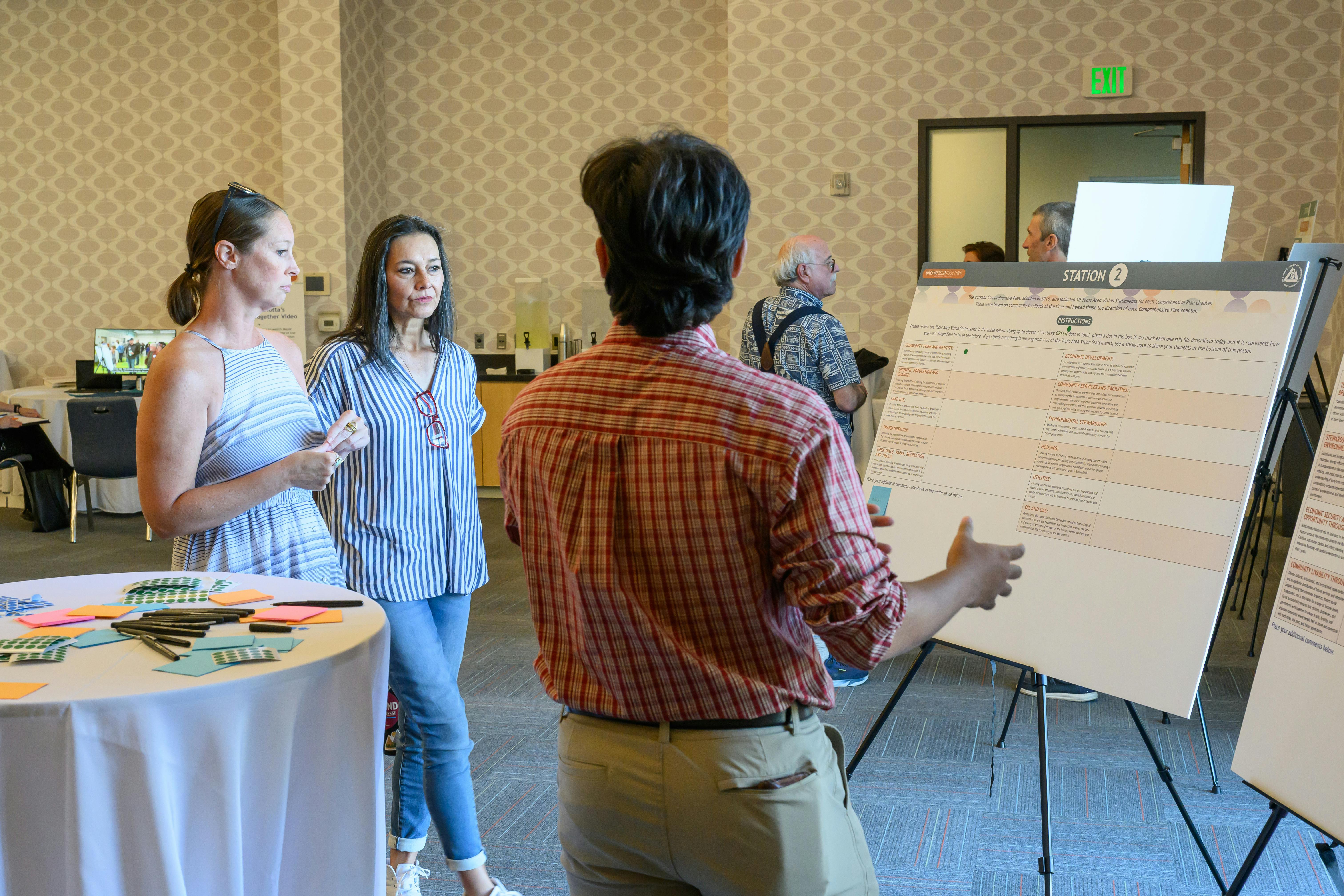 Photograph of a community engagement workshop where a facilitator in a red plaid shirt gestures toward a large informational poster. Two women listen as they stand near a table filled with pens, sticky notes, and green dot stickers for providing feedback.
