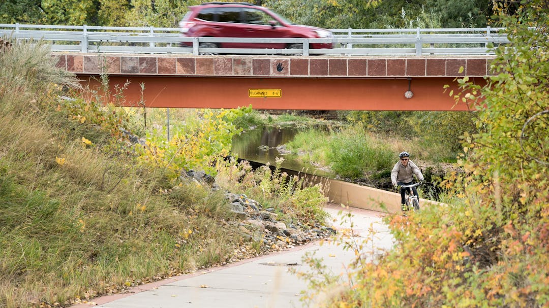 A cyclist is seen riding a riverside path under a bridge on which a car is passing.