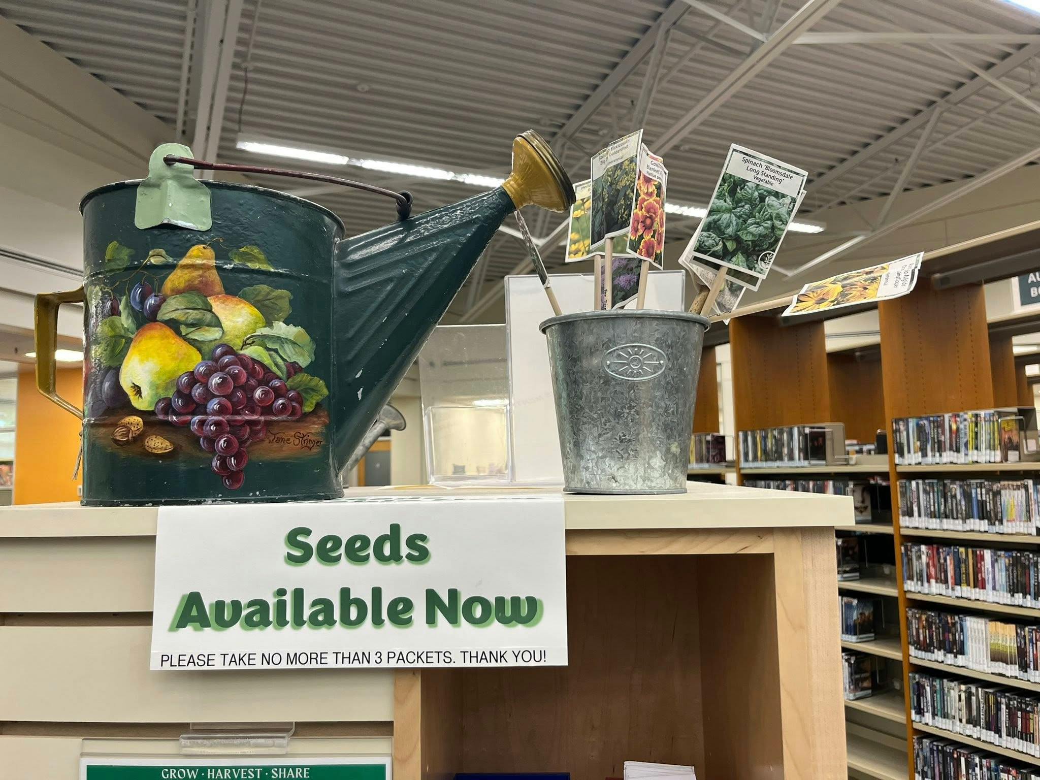 Watering can and seed packets with seeds available now sign