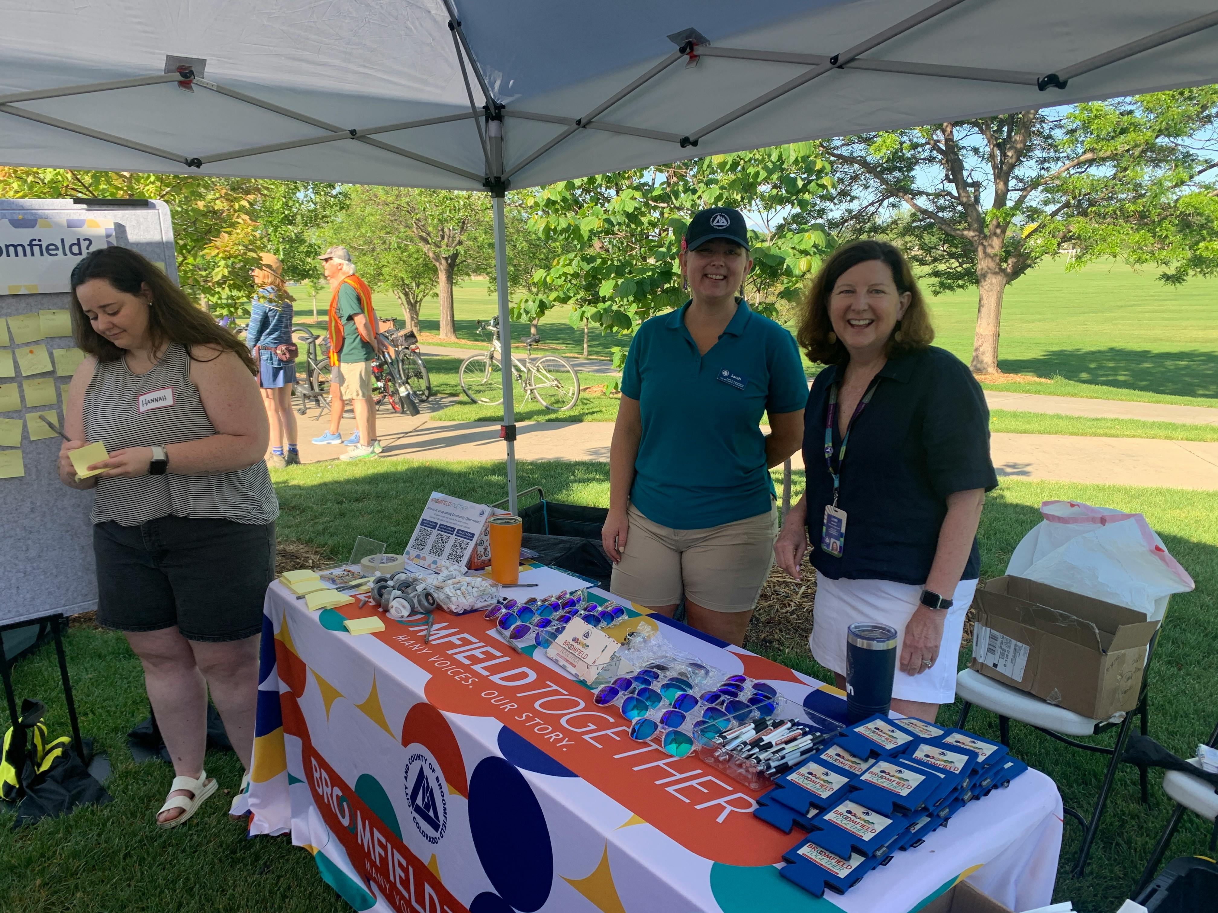 Two women stand behind a Broomfield Together table and smile for the camera. The table has project swag including sunglasses, koozies, bike bells and more.