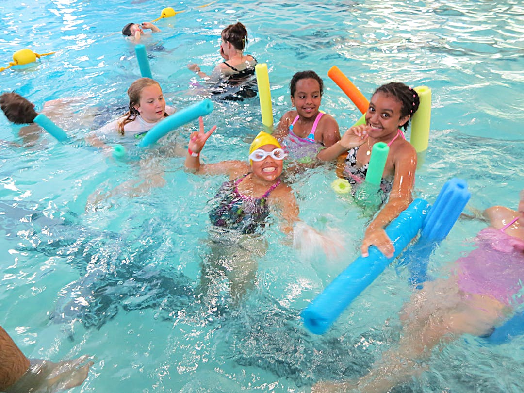 Kids swimming in an indoor swimming pool with pool noodles. 
