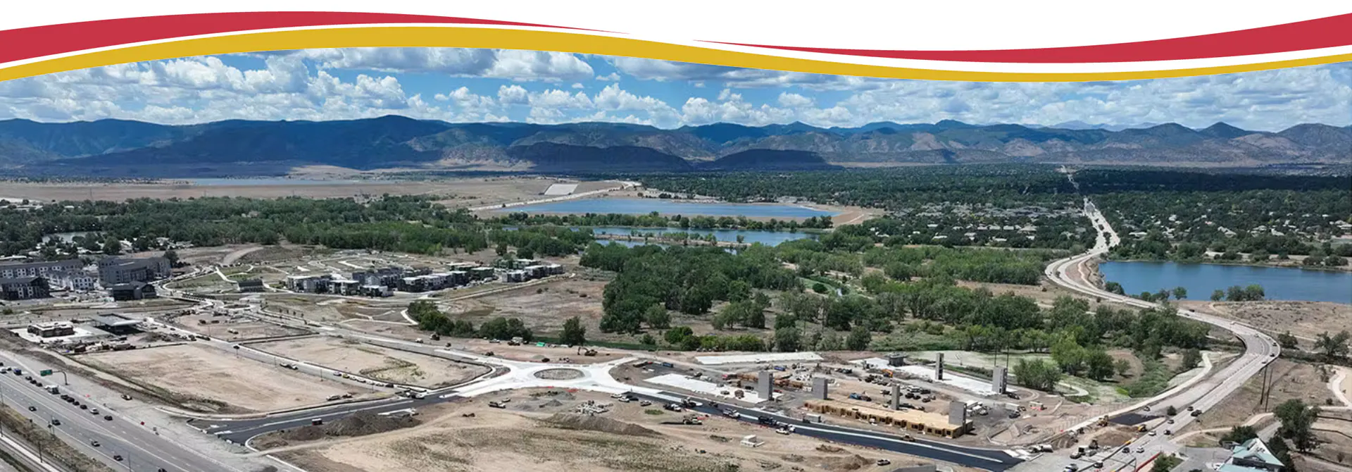 Aerial view of roundabout under construction surrounded by unfinished roads, buildings, mountains in distance.