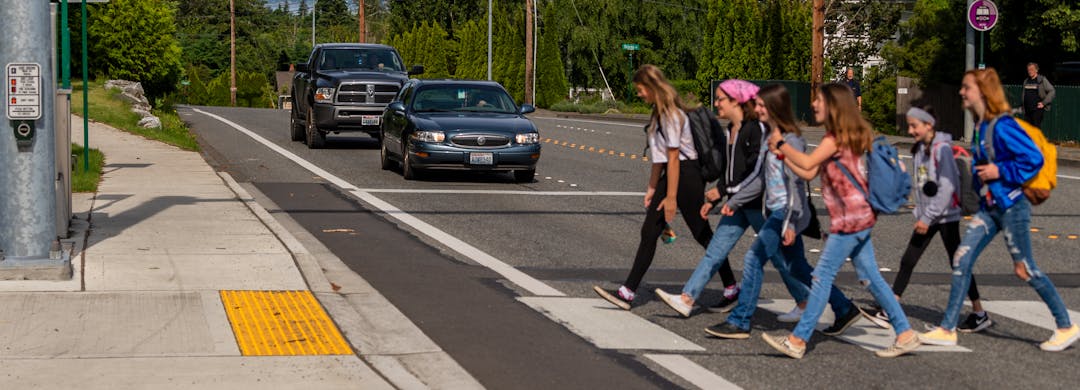 Six youth cross 4-lane road at designated crosswalk while car and truck are stopped to allow for safe crossing.