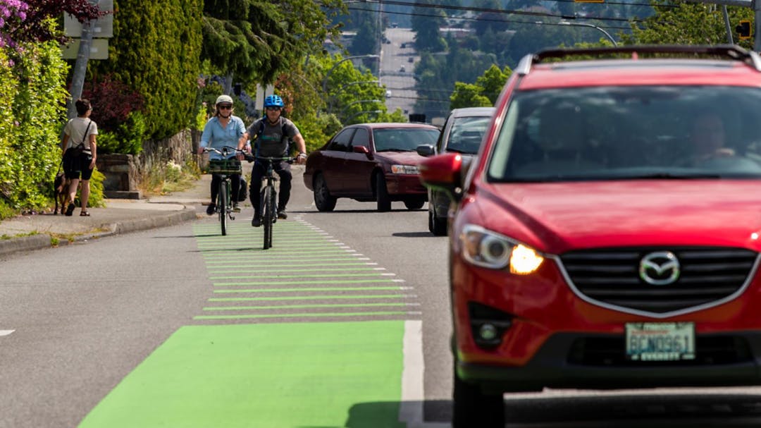 Two cyclist in green bike lane as they approach road intersection with red car waiting.