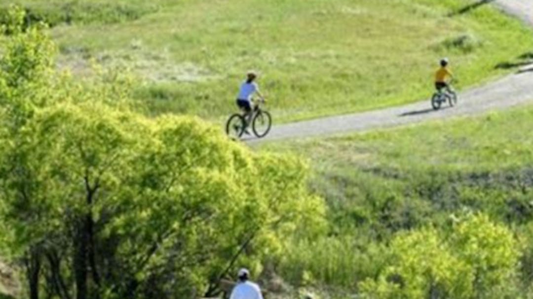 Image of people walking and riding bikes on a trail with bridge and green grass.
