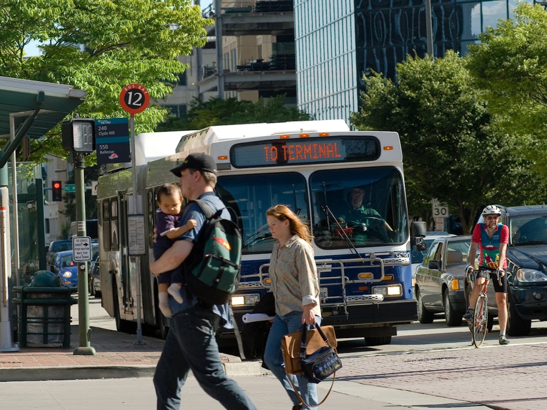 Two people walk in front of a bus, a person biking, and some cars waiting at a stop light.