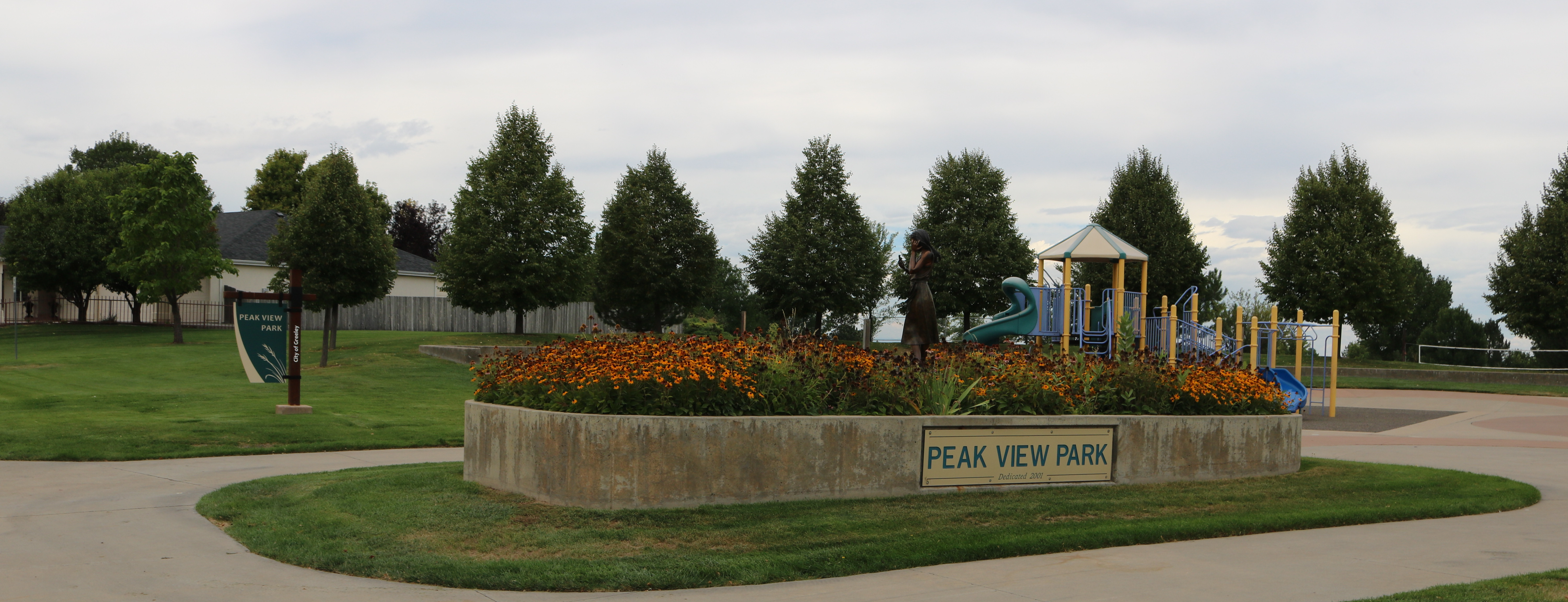 A park with green grass surrounds a multi-colored playgound with a concrete flowerbed with the name, "Peak View Park." 