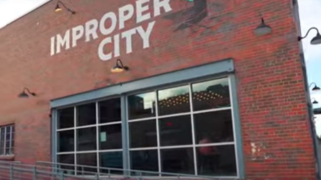 A brick building with a large, multi-pane window with the words "Improper City" painted in white above the window.