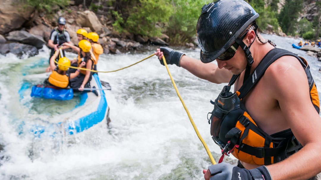 A man in a life vest holds a rope out to a raft full of people caught sideways in a rapid.