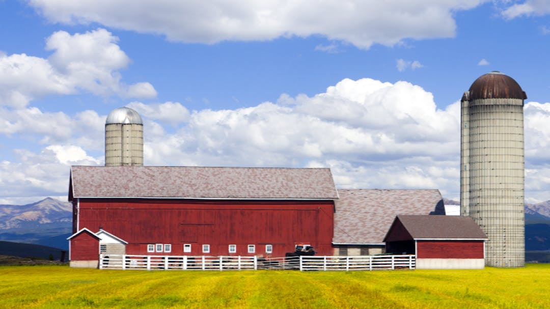 A large red barn in a field of yellow grass with a silo to the right and mountains behind it.
