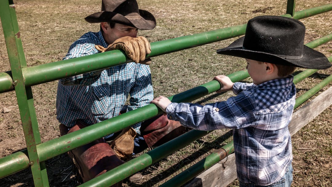 A cowboy in a blue shirt and cowboy hat kneels behind a green pole fence talking to a young boy also in western wear.