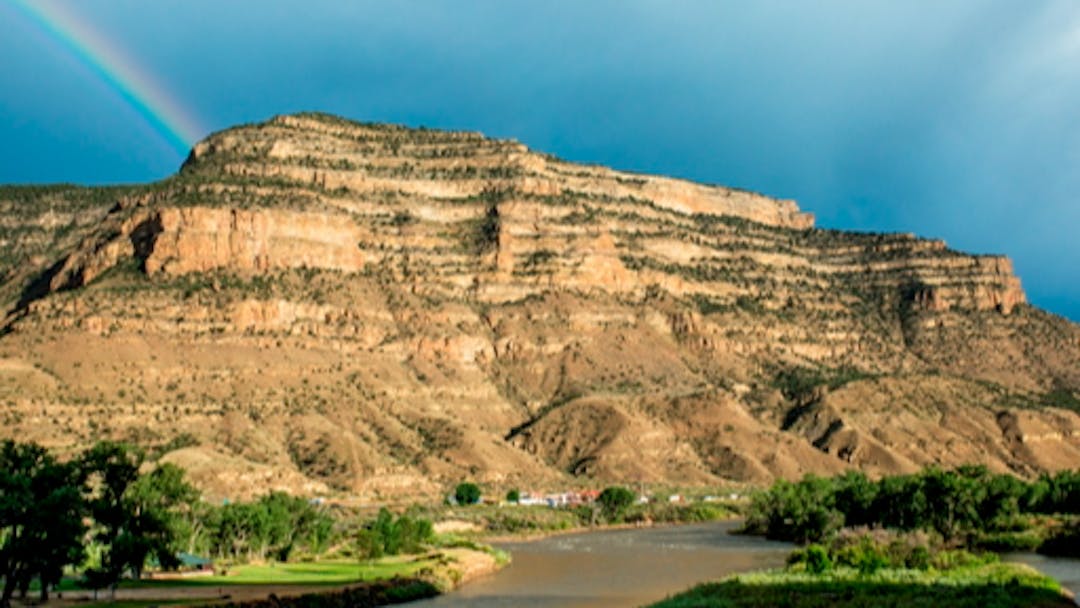 A cream-colored mesa sits behind a braided river with trees and a park on its banks.