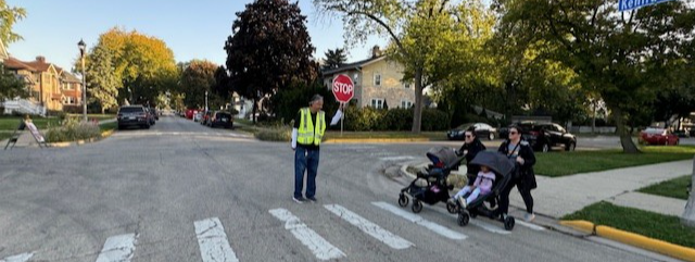 Two people walking with strollers as a crossing guard watches on.