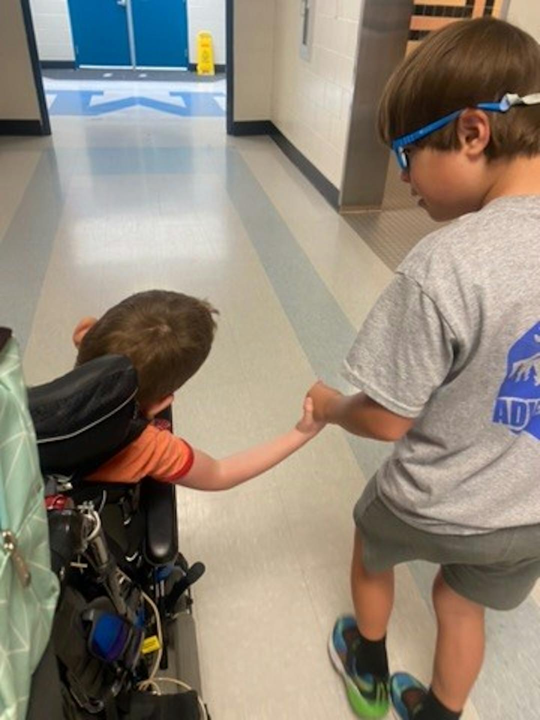 Shows a boy holding hands and helping another boy who is in a wheelchair.