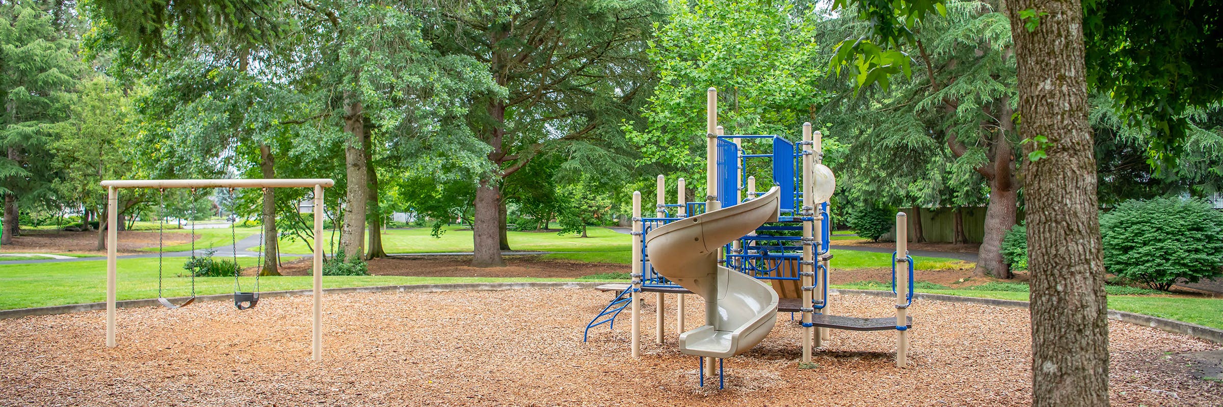 An old blue and tan metal play structure with slides and platforms surrounded by wood chips in a lush, green park. 
