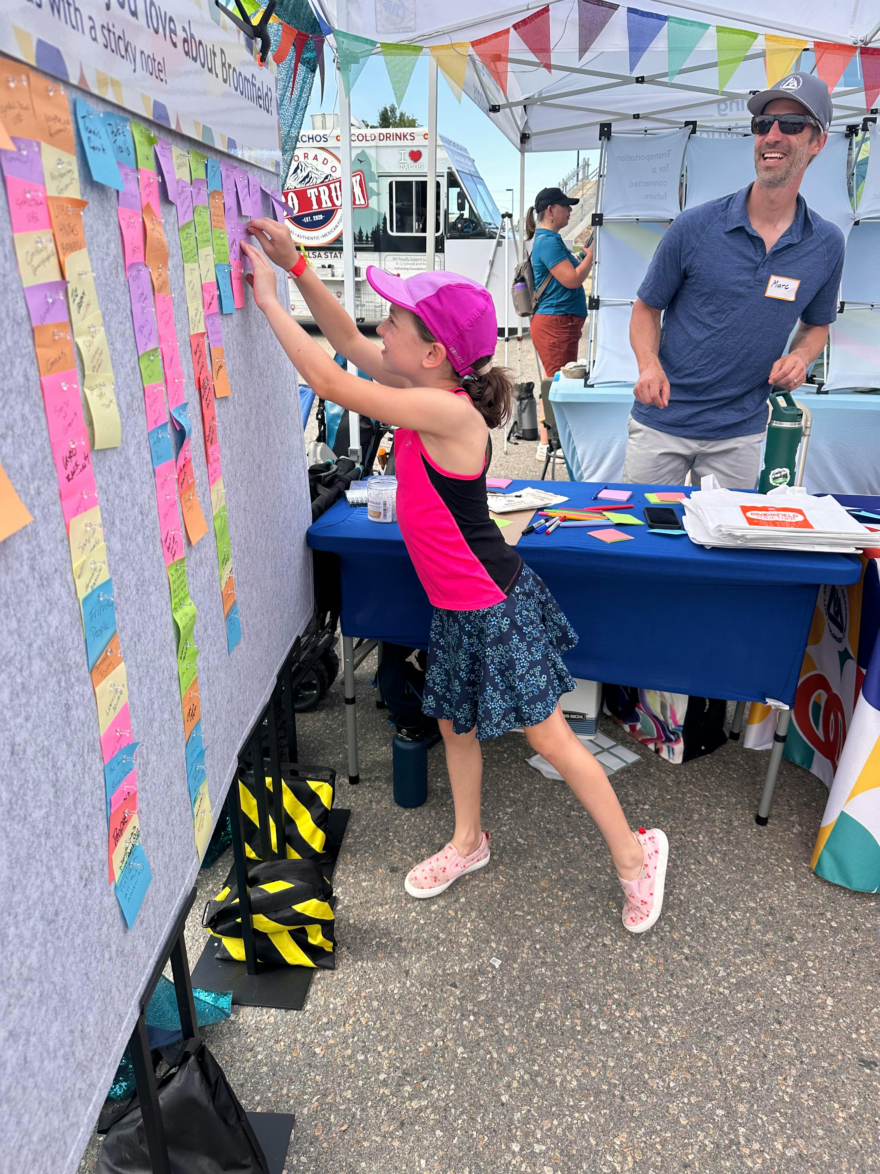 A young girl in a pink hat and tank top pins a sticky note to a large display board. The board is covered in numerous colorful handwritten notes. To the right, a man stands behind a table with event materials.