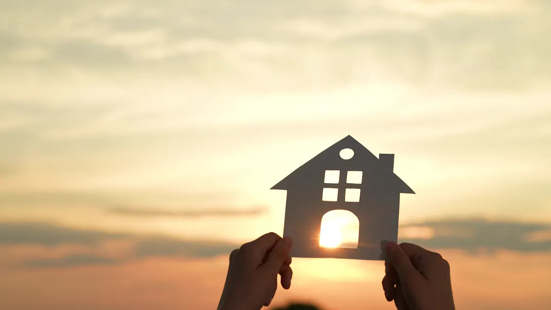 A person holds up a cutout of a home with the sun shining through in the background