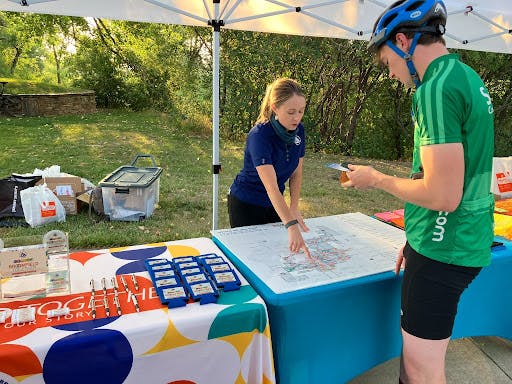 A woman in a blue polo shirt points to a map on a table under a white canopy tent, providing information to a man in a green cycling jersey and helmet. The scene takes place outdoors in a grassy park area with trees in the background.