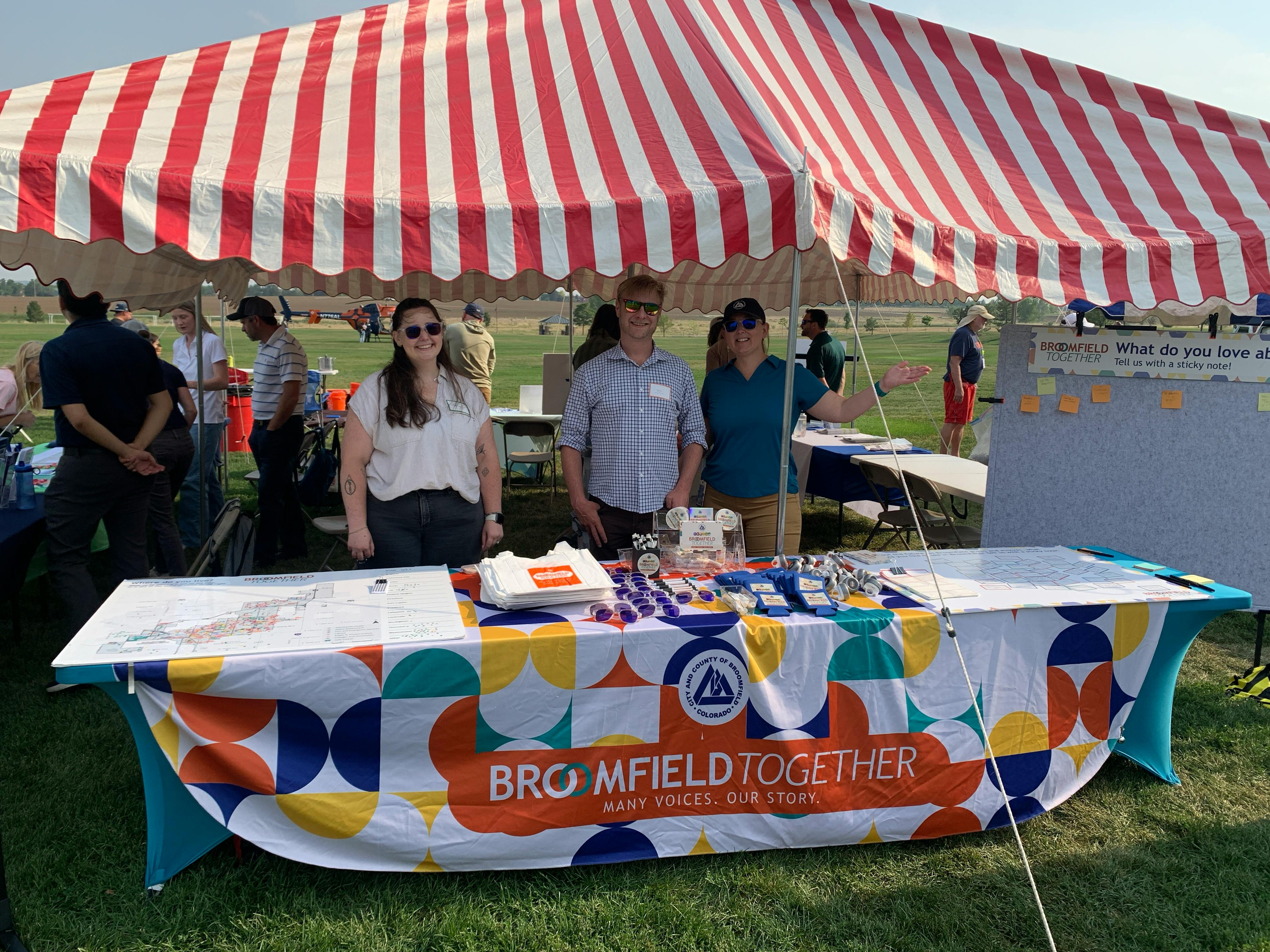 Photograph of three people standing behind a community engagement table under a red and white striped tent at an outdoor event. The table features a colorful tablecloth with the text "BROOMFIELD TOGETHER MANY VOICES. OUR STORY." and the CCOB logo.
