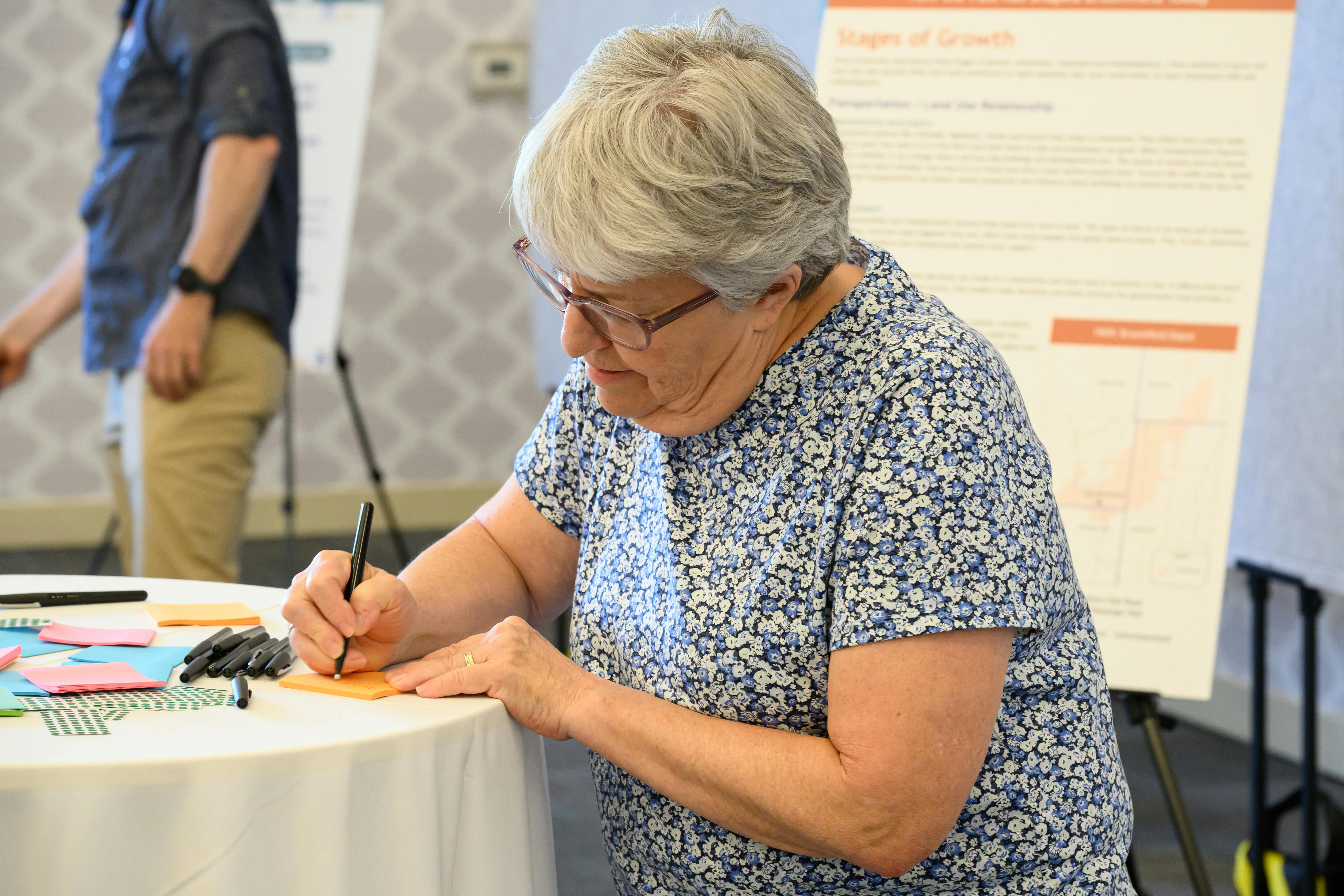 A woman with grey hair and glasses, wearing a blue floral shirt, writes on an orange sticky note at a table during a community workshop. In the background, a poster titled "Stages of Growth" is visible.