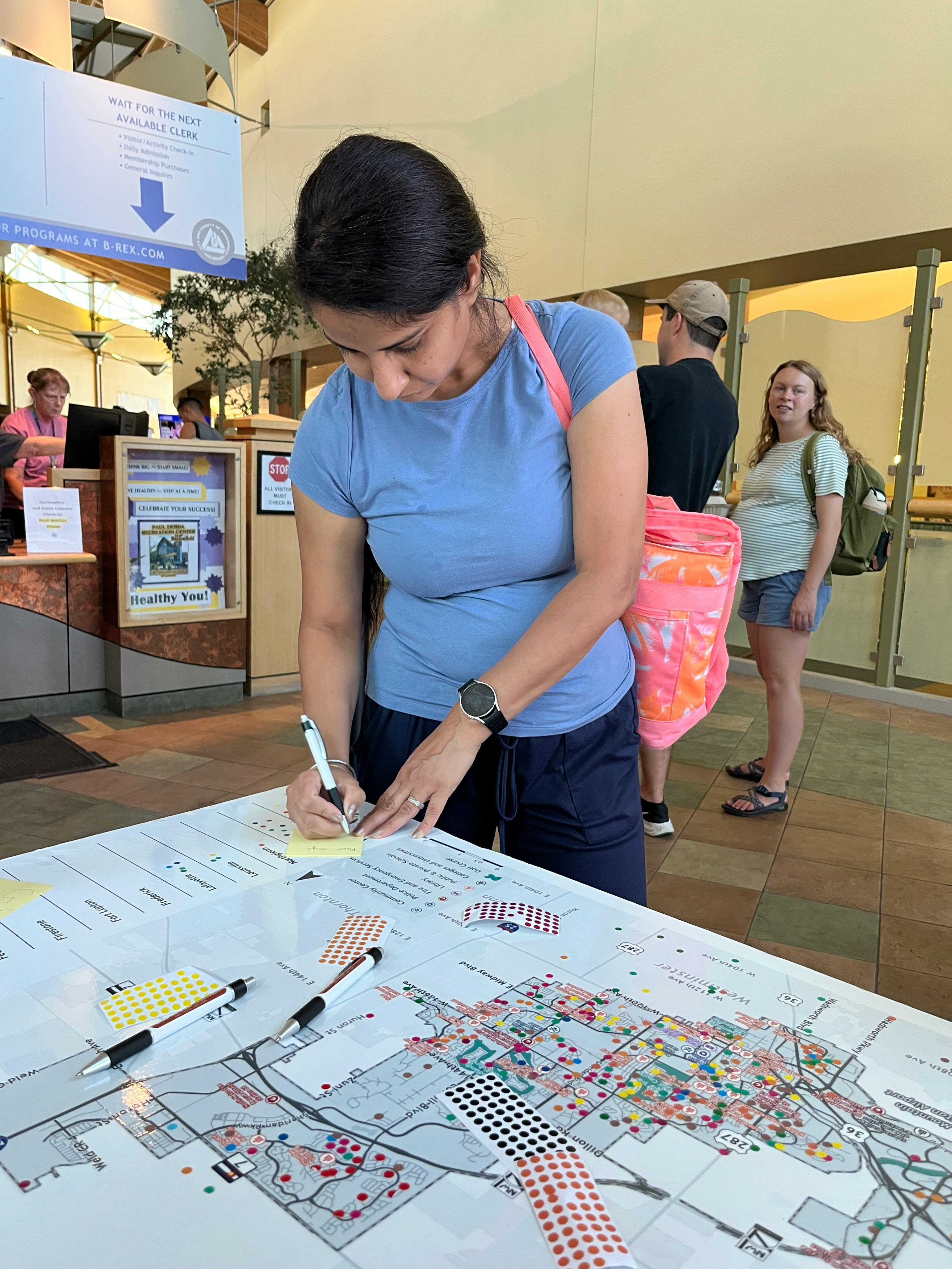 A woman in a blue t-shirt writes on a yellow sticky note placed on a large street map spread across a table. The map is marked with many small, colored dot stickers, indicating a community feedback activity.
