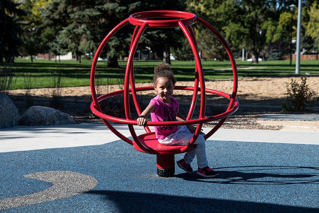 Young girl with brown skin and curly hair smiling while sitting inside playground equipment that looks like a large red metal ball.