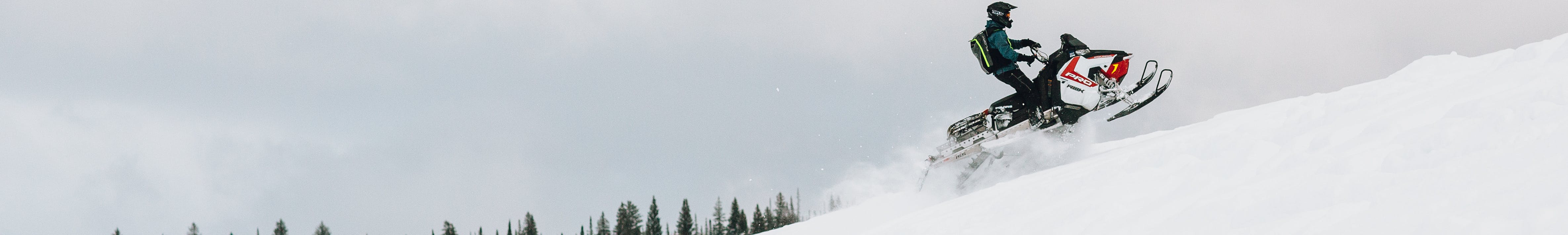 Snowmobile rider going up a snowy hill with trees in the background