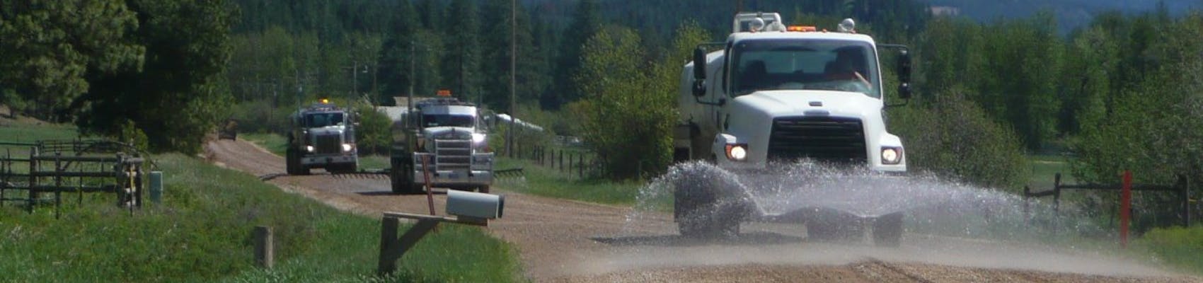 Dust abatement on rural County dirt road in summer. Shows three trucks.