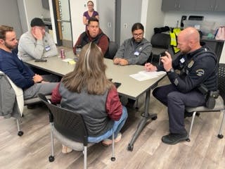 Employees sit around a table while a police officer in uniform takes notes